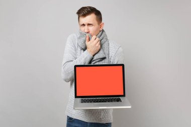 Boş boş ekran gri arka plan üzerinde izole ile dizüstü pc bilgisayar tutan eşarp ile ağız kapsayan gri süveterli genç adam. Sağlıklı yaşam, online tedavi danışmanlık, soğuk sezon kavramı