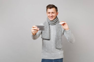 Gri süveterli, termometre, tutan eşarp genç adam gülümseyen selfie yaparken cep telefonu, video arama gri arka plan üzerinde izole yapma vurdu. Sağlık hasta hasta hastalık tedavileri, soğuk sezon kavramı