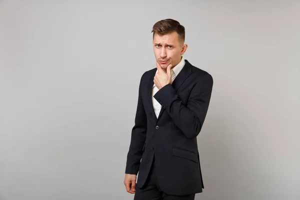Portrait of concerned young business man in classic black suit, shirt put hand prop up on chin isolated on grey background in studio. Achievement career wealth business concept. Mock up copy space