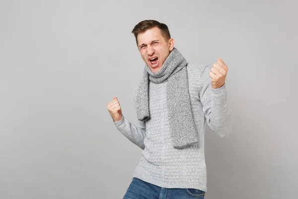 Joyful young man in gray sweater, scarf screaming, clenching fists like winner isolated on grey background. Healthy fashion lifestyle, people sincere emotions, cold season concept. Mock up copy space