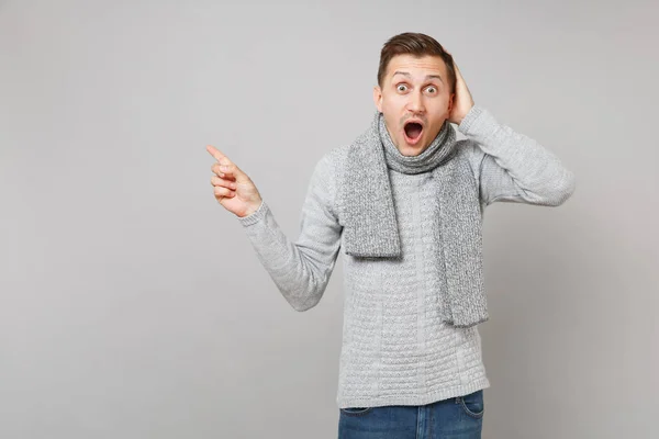 兴奋的年轻人穿着灰色毛衣 围巾把手放在头上 把食指指到一边 在灰色背景上张开嘴 健康的生活方式的人的情绪 寒冷的季节概念 — 图库照片
