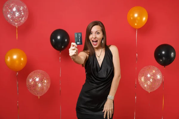 Mujer joven emocionada en pequeño vestido negro celebrando, mostrando la tarjeta de crédito en la cámara en globos de aire de fondo rojo. Día Internacional de la Mujer, Feliz Año Nuevo, concepto de fiesta de cumpleaños simulada . —  Fotos de Stock