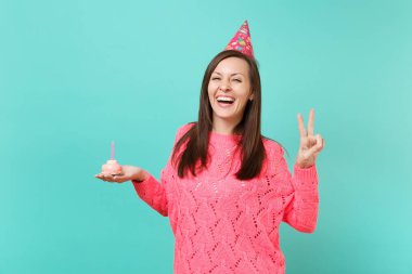 Gülümseyen örme pembe kazaklı genç kadın, doğum günü şapkası gösteren zafer işareti, tutun el kek ile mavi arka plan stüdyo portre üzerinde izole mum. İnsanlar yaşam tarzı kavramı. Kopyalama alanı alay