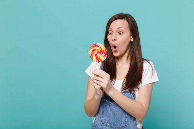 Denim giyim ağız geniş tutmak açık hayret genç kadın portresi, holding renkli yuvarlak lolipop Studio turkuaz mavi zemin üzerine izole. İnsanlar yaşam tarzı kavramı. Kopyalama alanı alay