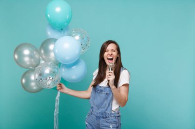 Denim giysiler içinde deli komik genç kadın şarkı mikrofon, kutlama ve izole mavi turkuaz arka plan üzerinde renkli hava balonları tutarak. Doğum günü tatil partisi, insanların duyguları kavramı