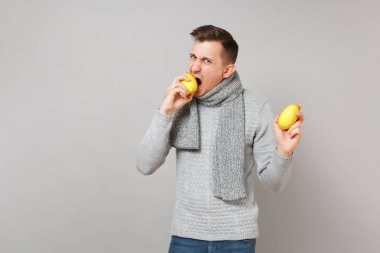 Gri süveterli holding, eşarp genç adam gri duvar arka planda, stüdyo portre izole limon ısırma. Sağlıklı moda yaşam tarzı, insanlar samimi duygular, soğuk sezon kavramı. Kopyalama alanı alay
