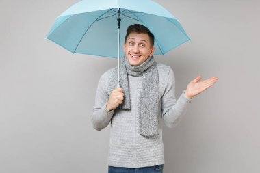 Hayret genç adam gri süveterli eşarp işaret ederek, gri arka plan üzerinde izole mavi şemsiye tutan el yayılıyor. Sağlıklı moda yaşam tarzı, insan duyguları, soğuk sezon kavramı. Kopyalama alanı alay