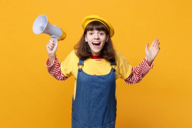 Heyecanlı kız genç Fransız bere, denim sundress holding megafon, sarı duvar arka planda Studio izole eller yayılıyor. İnsanlar samimi duygular, yaşam kavramı. Kopyalama alanı alay