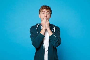 Ayakta, mavi duvar arka planda Studio izole ellerle ağız kapsayan rahat kıyafetler şok hayran genç adam portresi. İnsanlar samimi duygular, yaşam kavramı. Kopyalama alanı alay