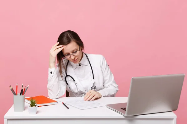 Tired sad female doctor sits at desk work on computer with medical document in hospital isolated on pastel pink wall background. Woman in medical gown glasses stethoscope. Healthcare medicine concept