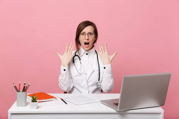 Tired mad female doctor sits at desk work on computer with medical document in hospital isolated on pastel pink wall background. Woman in medical gown glasses stethoscope. Healthcare medicine concept