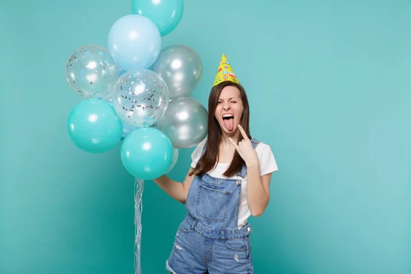 Crazy girl in birthday hat showing tongue horns up heavy metal rock sign celebrating, hold colorful air balloons isolated on blue turquoise background. Birthday holiday party, people emotions concept