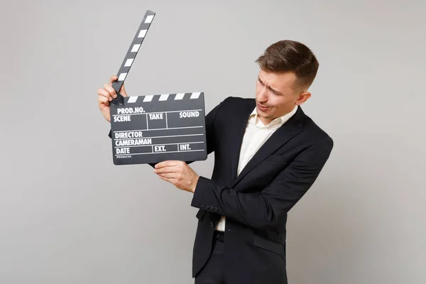 Young business man in classic black suit looking on classic black film making clapperboard in hands isolated on grey wall background. Achievement career wealth business concept. Mock up copy space