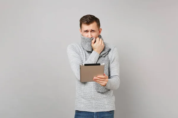 Young man in sweater covering mouth with scarf, coughing using tablet pc computer isolated on grey background. Healthy lifestyle, online treatment consulting, cold season concept. Mock up copy space