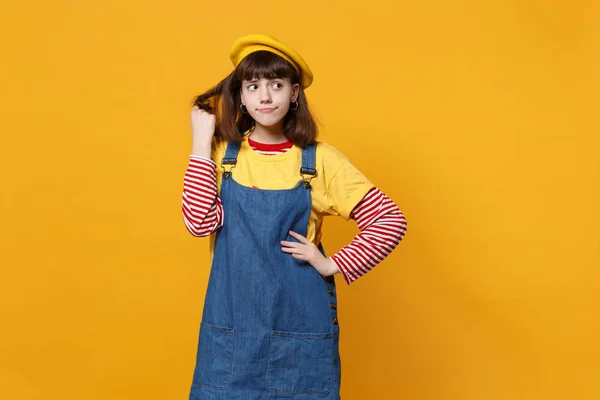 Puzzled pensive girl teenager in french beret, denim sundress holding hair, looking aside isolated on yellow wall background in studio. People sincere emotions, lifestyle concept. Mock up copy space