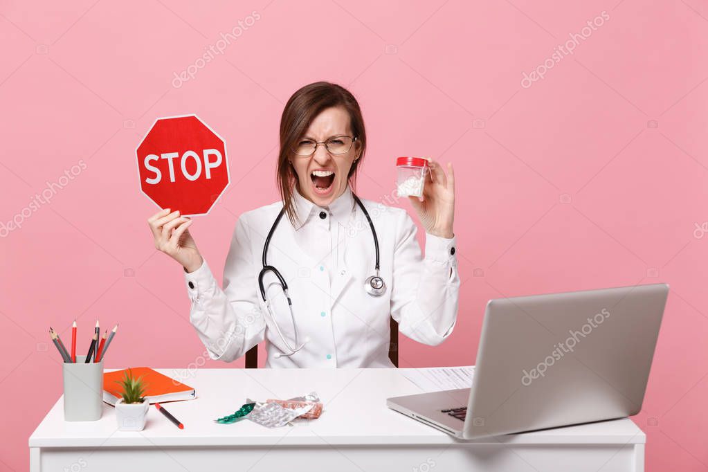 Female doctor sit at desk work on computer with medical document hold pills in hospital isolated on pastel pink background. Woman in medical gown glasses stethoscope. Healthcare medicine concept.