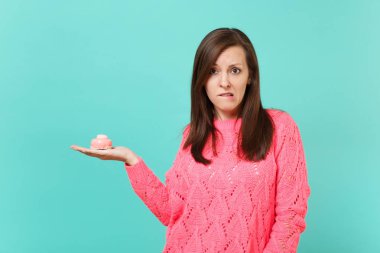 Örgü pembe kazak elinde tutan pasta ve mavi turkuaz duvar arka planda, stüdyo portre izole dudak ısırma rahatsız genç kadın şaşkın. İnsanlar yaşam tarzı kavramı. Kopyalama alanı alay