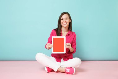 Tam uzunlukta gül gömlek bluz, kadın portre, beyaz pantolon yere oturma parlak pembe mavi pastel duvar arka plan studio izole tablet pc tutun. Moda yaşam tarzı kavramı. Kopyalama alanı alay