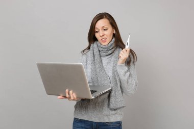 Söz konusu genç kadın gri süveterli dizüstü pc bilgisayar üzerinde çalışıyor gri duvar arka plan üzerinde izole termometre tutan eşarp. Sağlıklı yaşam, online tedavi danışmanlık, soğuk sezon kavramı