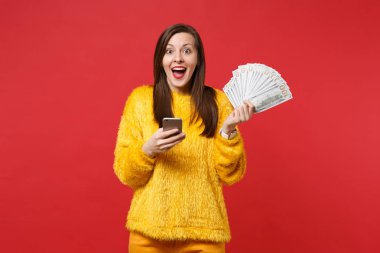 Cep telefonu, fan Doları banknot, kırmızı zemin üzerine izole para para para tutan sarı kürk kazaklı şok genç kadın. İnsanlar samimi duygular, yaşam kavramı. Kopyalama alanı alay