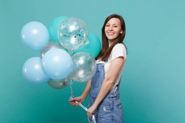 Çok gülümseyen genç mutlu kadın kutluyor ve izole mavi turkuaz duvar arka plan üzerinde renkli hava balonları tutan denim giysileri portresi. Doğum günü tatil partisi, insanların duyguları kavramı