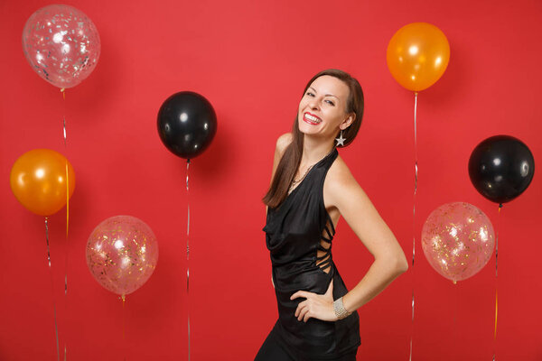 Smiling young girl in little black dress celebrating standing with arms akimbo on bright red background air balloons. St. Valentine's, Women's Day Happy New Year birthday mockup holiday party concept
