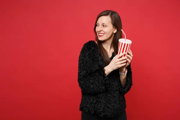 微笑的年轻女子穿着黑色皮毛衣看着一边 拿着塑料杯可乐或苏打水隔离在明亮的红色背景在工作室 人们真诚的情感 生活方式的理念 模拟复制空间 — 图库照片