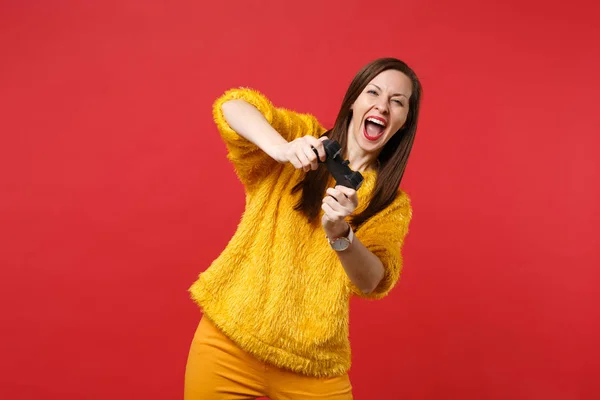 Portrait of funny laughing young woman in yellow fur sweater playing video game with joystick isolated on bright red wall background. People sincere emotions, lifestyle concept. Mock up copy space