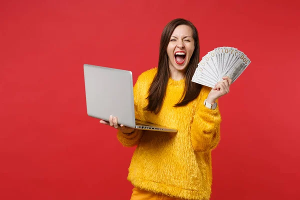 Crazy young woman in fur sweater screaming, holding laptop pc computer, fan of money in dollar banknotes, cash money isolated on red background. People emotions, lifestyle concept. Mock up copy space