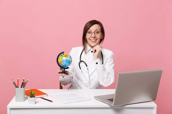 Female doctor sit at desk work on computer with medical document hold globe in hospital isolated on pastel pink wall background. Woman in medical gown glasses stethoscope. Healthcare medicine concept