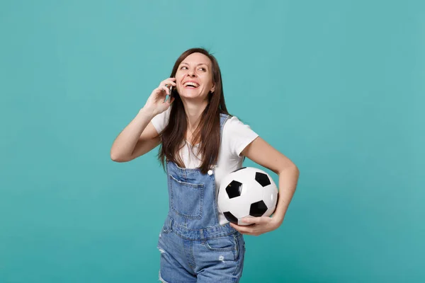 Laughing young woman football fan with soccer ball talking on mobile phone, conducting pleasant conversation isolated on blue turquoise wall background. People emotions, sport family leisure concept