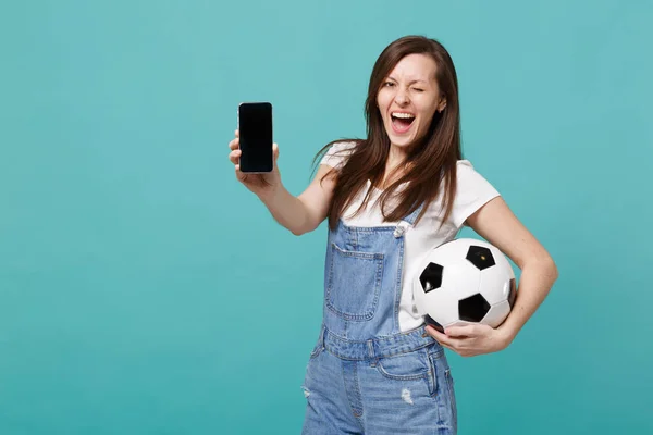Cheerful blinking woman football fan holding soccer ball, mobile phone with blank empty screen isolated on blue turquoise background. People emotions, sport family leisure concept. Mock up copy space
