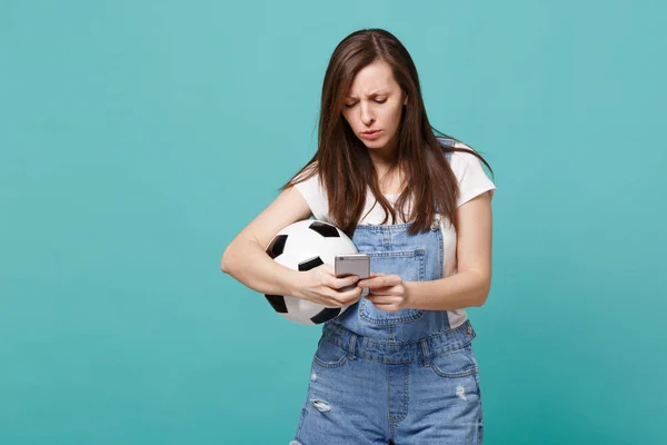 Puzzled young woman football fan with soccer ball using mobile phone, typing sms message isolated on blue turquoise wall background. People emotions sport family leisure concept. Mock up copy space