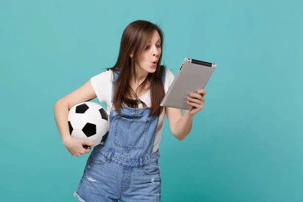 Amazed young woman football fan holding soccer ball, using tablet pc computer isolated on blue turquoise wall background. People emotions, sport family leisure lifestyle concept. Mock up copy space