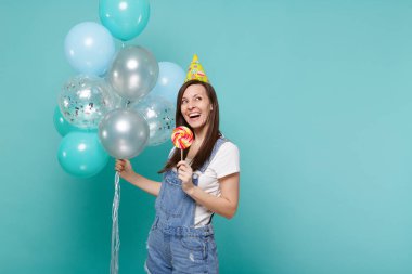 Ararken, lolipop tutan doğum günü şapkalı thinkful genç kadın izole mavi turkuaz arka plan üzerinde renkli hava balonları ile kutluyor. Doğum günü tatil partisi, insanların duyguları kavramı