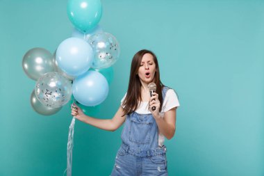 Denim giysiler içinde söz konusu genç kadın şarkı mikrofon, kutlama, izole mavi turkuaz duvar arka plan üzerinde renkli hava balonları bekletme. Doğum günü tatil partisi, insanların duyguları kavramı