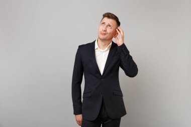 Dalgın şaşkın genç iş adamı klasik siyah takım elbise, ararken, Studio gri arka plan üzerinde izole baş el koyarak gömlek. Başarı kariyer servet iş kavramı. Kopyalama alanı alay