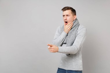 Genç adam gri kazak, eşarp görünümlü, işaret parmağı bir kenara ve gri arka plan üzerinde izole çene üzerinde yukarı koymak el pervane hayran. Sağlıklı yaşam, insanlar samimi duygular, soğuk sezon kavramı