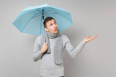 Şaşkın genç adam gri süveterli işaret eden bir ele bir yana, gri arka plan üzerinde izole mavi şemsiye tutan eşarp. Sağlıklı moda yaşam tarzı, insan duyguları, soğuk sezon kavramı. Kopyalama alanı alay