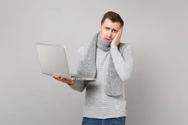 Kazaklı, yorgun genç adam eşarp dizüstü pc bilgisayar, gri arka plan üzerinde izole yanağına el koymak üzerinde çalışıyor. Sağlıklı yaşam, online tedavi danışmanlık, soğuk sezon kavramı. Kopyalama alanı alay