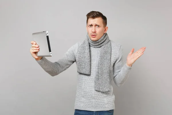 Puzzled young man in gray sweater, scarf making video call with tablet pc computer, spreading hands isolated on grey background. Healthy lifestyle, online treatment consulting, cold season concept