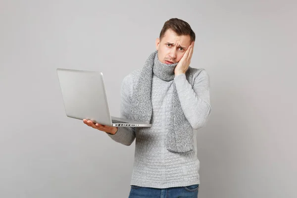 Tired young man in sweater, scarf working on laptop pc computer, put hand on cheek isolated on grey background. Healthy lifestyle, online treatment consulting, cold season concept. Mock up copy space