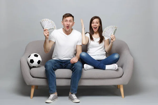 Excited couple woman man football fans cheer up support favorite team holding fan of money in dollar banknotes, cash money spreading hands isolated on grey background. Sport family lifestyle concept