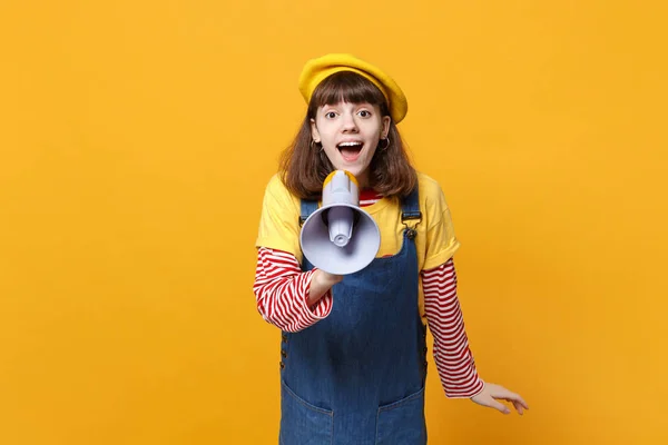 Portrait of excited cute girl teenager in french beret, denim sundress scream in megaphone isolated on yellow wall background in studio. People sincere emotions, lifestyle concept. Mock up copy space