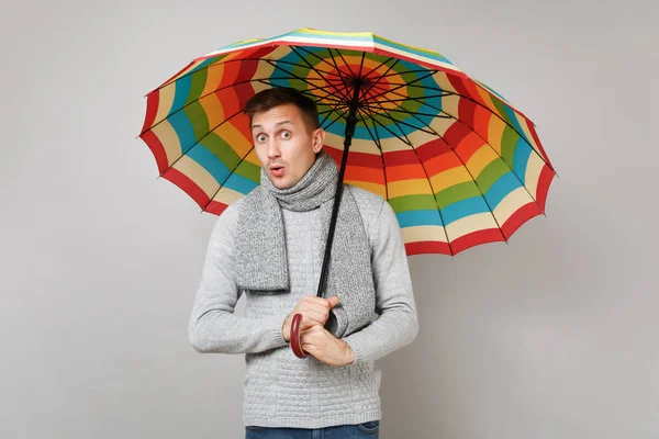 Σοκαρισμένος νεαρός άνδρας σε γκρι πουλόβερ, κασκόλ κρατώντας πολύχρωμες ομπρέλες — Φωτογραφία Αρχείου