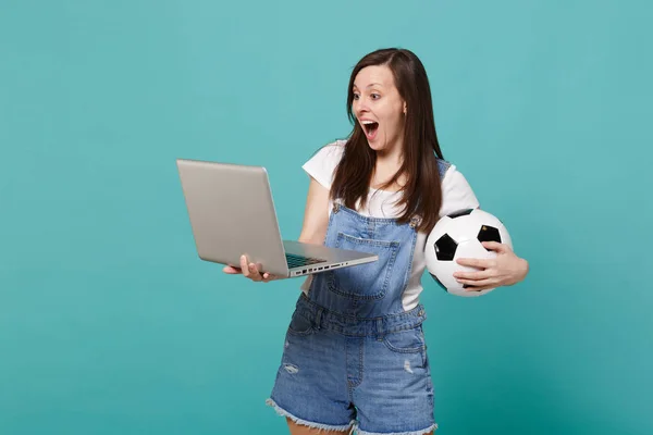 Excited young woman football fan holding soccer ball, using lapt