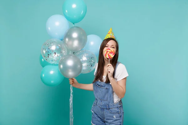 Funny young woman in birthday hat covering mouth with round loll