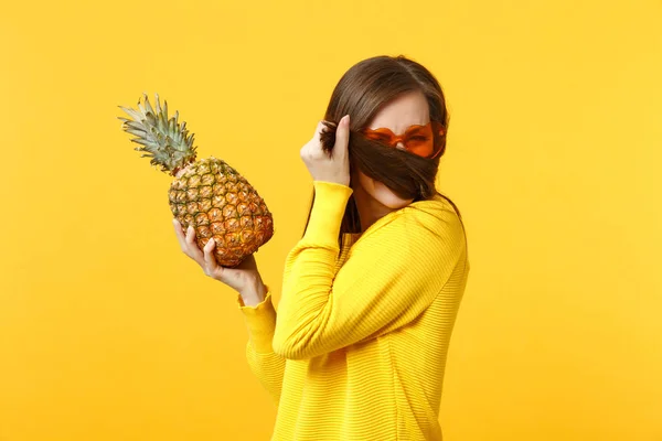 Jovencita divertida en gafas de corazón cubriendo la cara con pelo, sosteniendo fruta fresca de piña madura aislada sobre fondo amarillo anaranjado. Gente estilo de vida vívido, relajarse concepto de vacaciones. Simular espacio de copia . —  Fotos de Stock