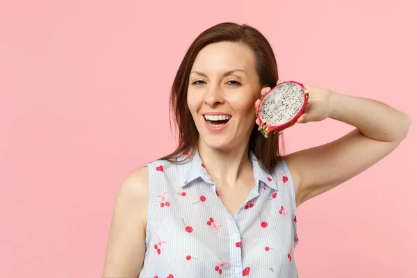 Cheerful young woman in summer clothes holding half of fresh ripe pitahaya dragon fruit isolated on pink pastel background in studio. People vivid lifestyle relax vacation concept. Mock up copy space.
