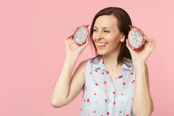 Joyful young woman in summer clothes looking aside, hold half of fresh ripe pitahaya dragon fruit isolated on pink pastel background. People vivid lifestyle relax vacation concept. Mock up copy space.